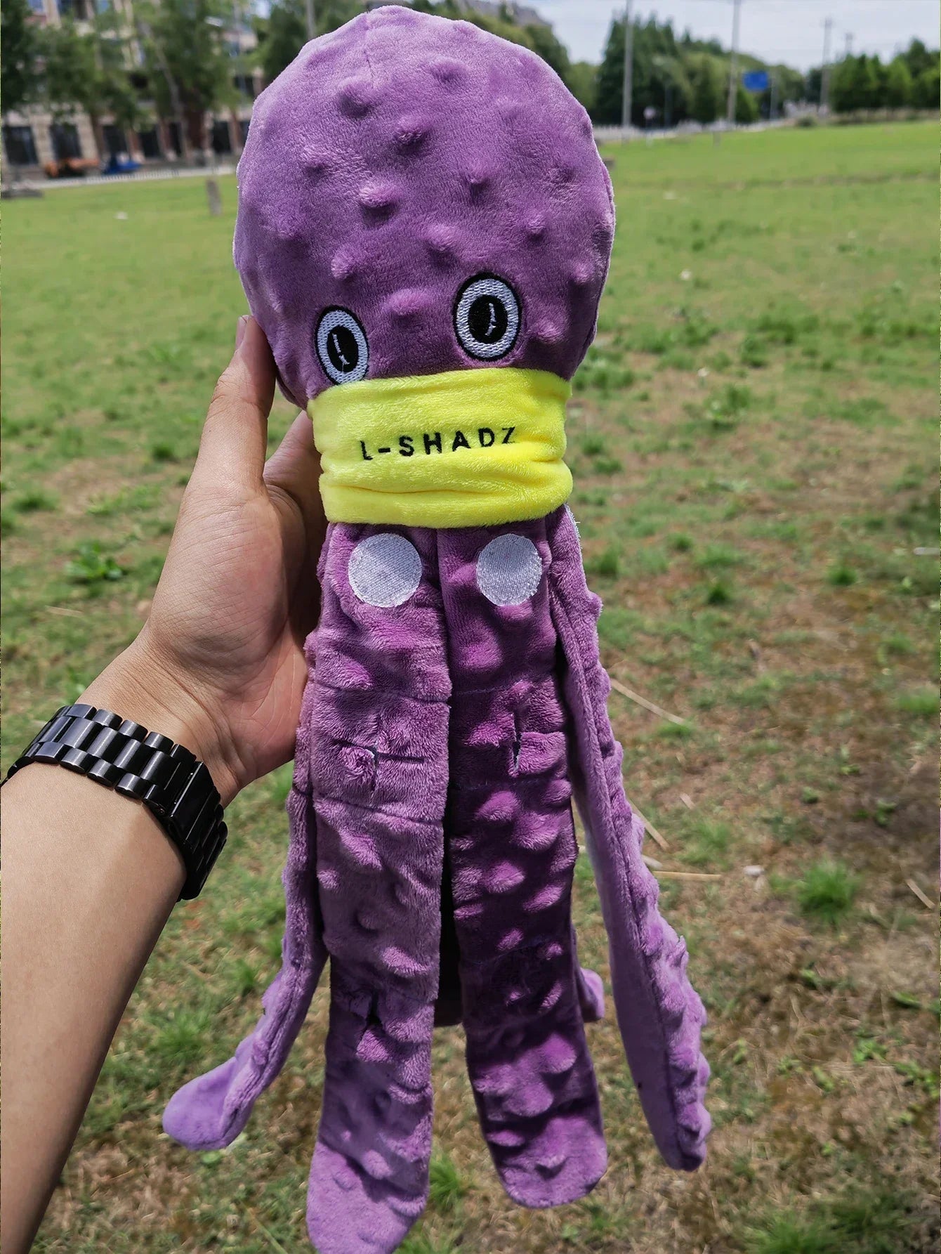 Purple plush octopus toy with a yellow bandana labeled 'L-Shadz' held in a hand outdoors.